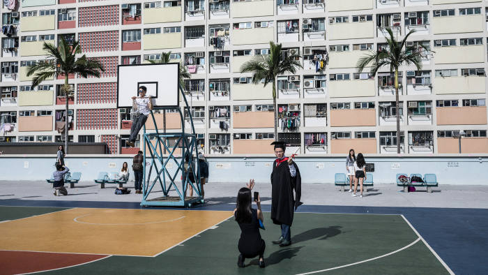 A student graduate poses for a photograph as a man hangs from a basketball hoop in front of residential homes at a public housing estate in Hong Kong on April 1, 2017. / AFP PHOTO / DALE DE LA REY (Photo credit should read DALE DE LA REY/AFP/Getty Images)