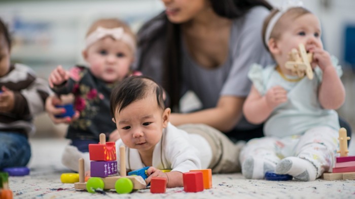 A multi-ethnic group of babies are indoors in a daycare center. They are wearing casual clothing. They are playing with toys along with their babysitter. One baby is crawling on the floor and playing with a car in the foreground.