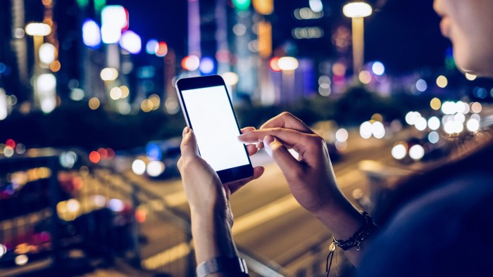 Portrait of woman text messaging on smartphone in city street at night, with busy traffic scene and illuminated skyscrapers in the background.