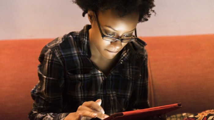 This is a horizontal, close up, color, royalty free stock photograph of an intelligent Black American woman in her 20s browsing the internet with a digital tablet while sitting on the couch in her Brooklyn apartment in New York City. As she scrolls through web pages online the glowing screen illuminates her face. Photographed with a Nikon D800 DSLR camera.