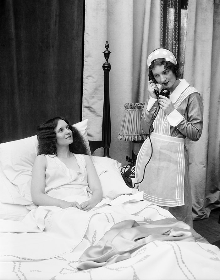 Two women in bedroom: one dressed as a maid on the phone, the other in bed smiling up at the maid