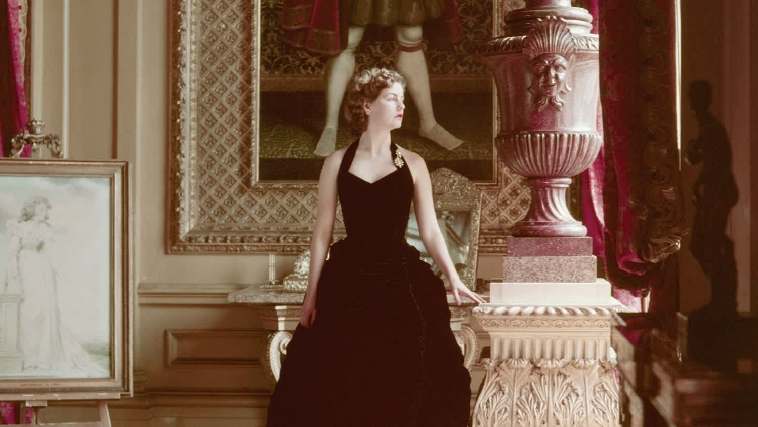 An elegant woman in a black evening gown stands poised in a lroom with richly patterned walls, beside a large framed portrait of a regal figure, with a white dog sleeping in a beam of sunlight on the carpet