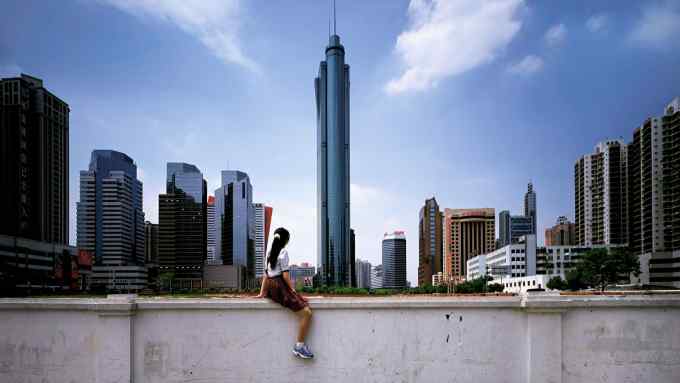 A girl or young woman with long black hair, white blouse, skirt and trainers sits on a wall facing towards a cityscape of tower blocks and a gleaming skyscraper