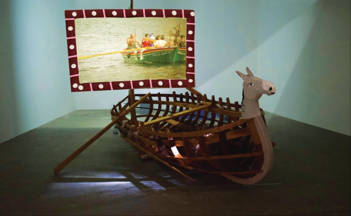 In a gallery setting, a wooden rowing boat with a horse mask on its prow is placed in front of a screen showing a video of people on a rowing boat