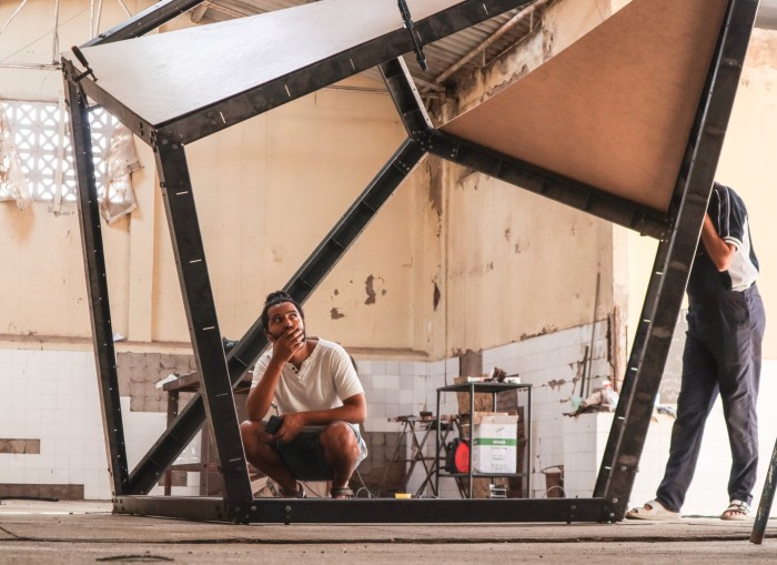 A bearded man crouches inside of the metal frame of an abstract sculpture in a studio