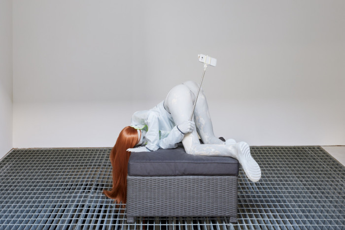 A white mannequin of a young woman crouches down on a cubic grey sofa with its back up in the air while holding a selfie stick complete with phone in its left hand