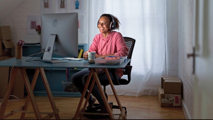 A young woman in a pink shirt and blue jeans sits at her desk as she works from home