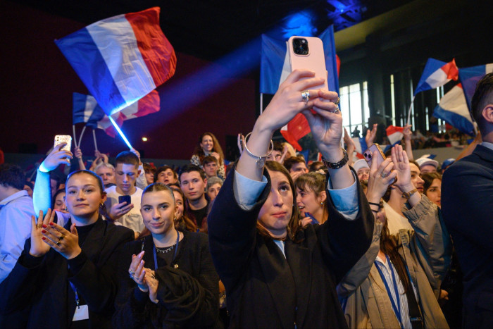 Young RN supporters waving French flags hold up their phones to photograph speakers at a party rally