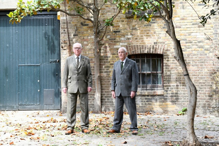 Two middle-aged men dressed in formal suits stand in in front of a brick house and garage