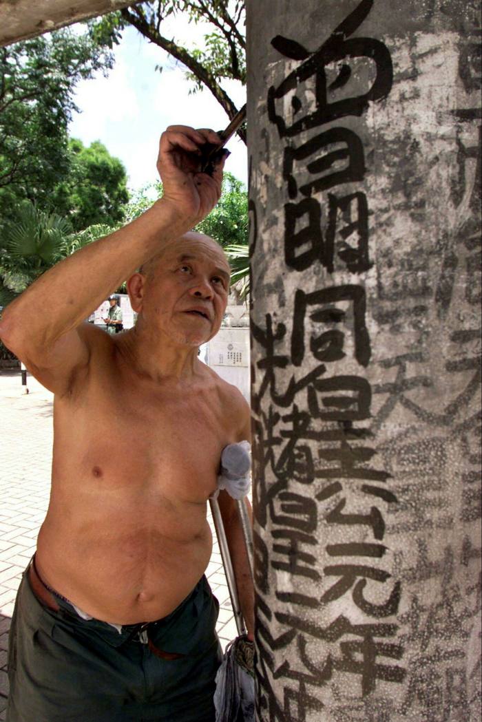 A shirtless older man paints Chinese writing in black ink onto a bus stop