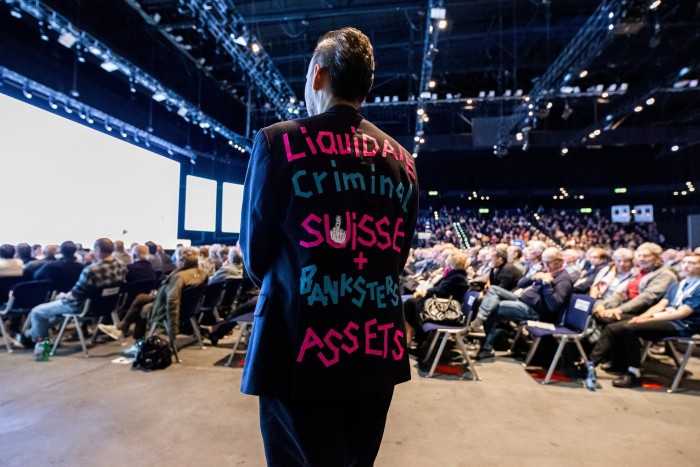 A man at a Credit Suisse shareholder meeting wearing a blazer saying:  Liquidate criminal Suisse 