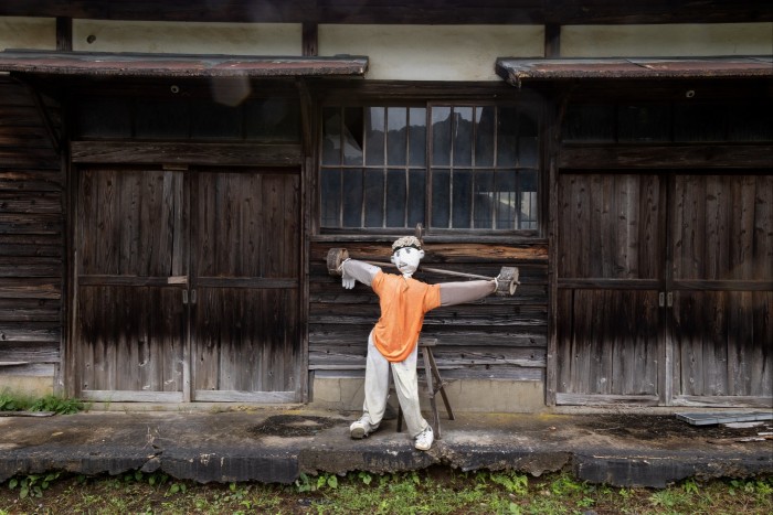A stuffed figure leans next to a wooden building, arms outstretched as if weight lifting