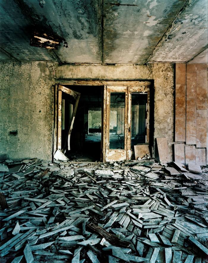 In an abandoned building, grey pieces of a wooden floor are piled up irregularly in front of a rusty door