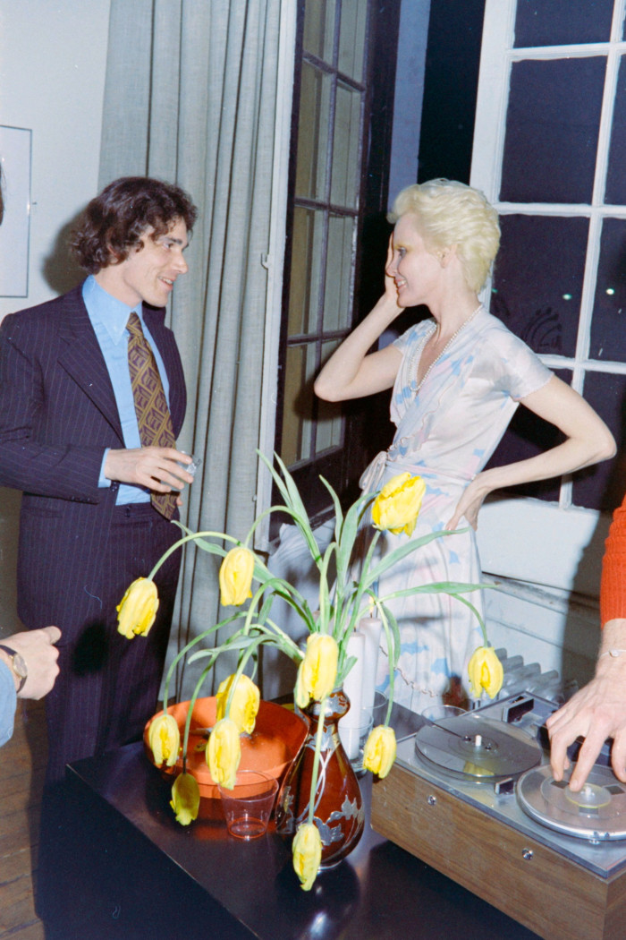 A man in a suit stands holding a drink and talking to a woman in a loft studio, with a vase of flowers on a table in front of them
