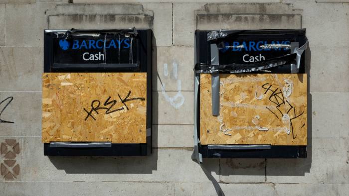 Two cashpoints side by side on a bank wall, boarded up with chipboard and partly repaired with tape. Graffiti scrawled across them and on the wall