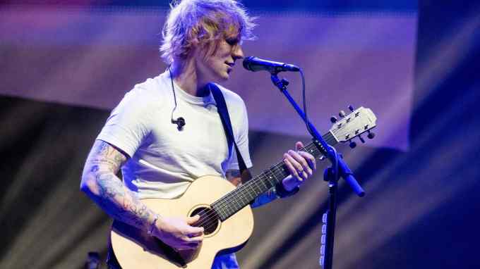 Ed Sheeran, with messy ginger hair and in a white T-shirt, is playing the guitar and singing into a microphone