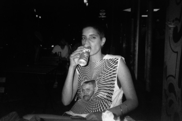 A black and white photo of a woman drinking a Sprite, wearing an abstract-patterned vest