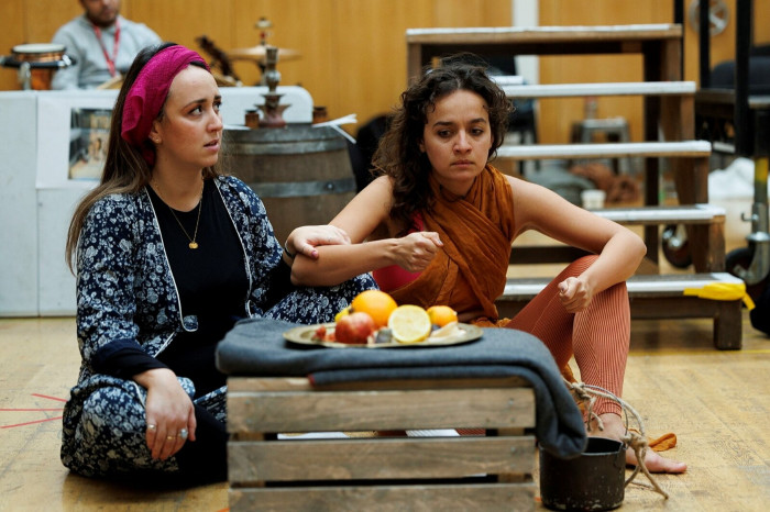 Two women sit cross-legged at a makeshift table formed by a wooden crate, as they ponder sliced fruit on a plate