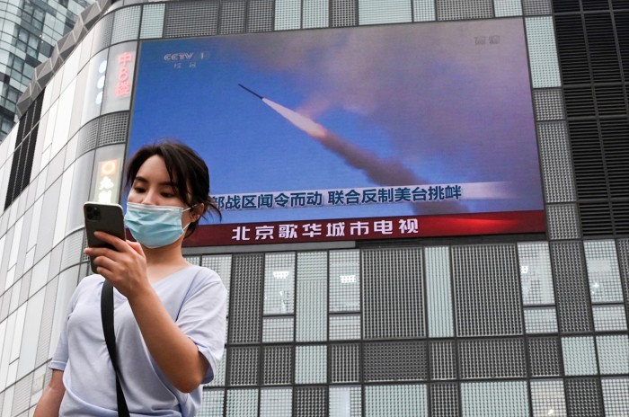 A woman uses her mobile phone as she walks in front of a large screen showing a news broadcast about China’s military exercises encircling Taiwan