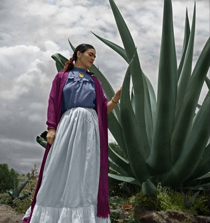 Frida Kahlo stands in front of a large aloe plant