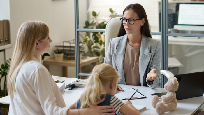 A mother talks to another woman in glasses who is behind a desk. The mother sits beside her child who seems to be writing down some notes