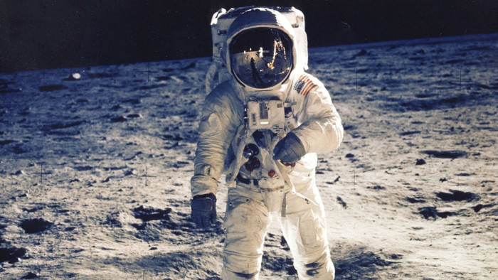 A historic photograph of an astronaut in a white space suit standing on the moon’s surface, with the lunar lander reflected in the visor of the helmet
