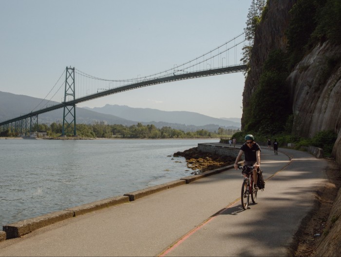 Lions Gate Bridge, Vancouver, as seen on a sunny day from the Stanley Park Seawall skirting the harbour. A male cyclist is in the foreground