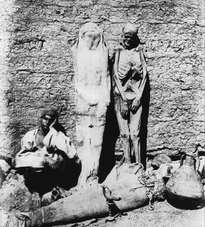 A man sits on the ground, his head leaning against his hand, beside two standing mummies, one still in its shroud, the other just a skeleton