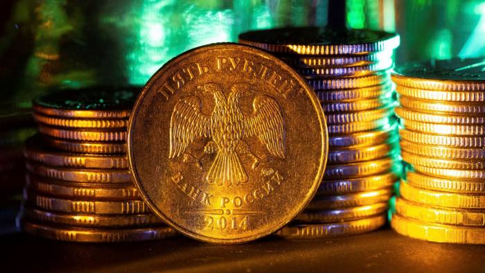 The Russian imperial double-headed eagle coat of arms, the emblem of Russia’s central bank, sits on a five rouble coin, standing on it’s edge against piles of rouble coins