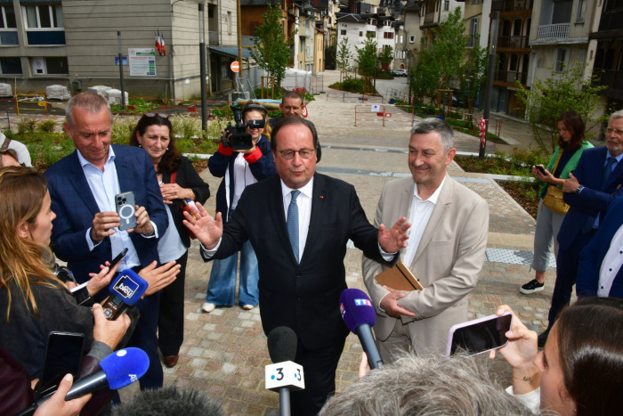 A man in a suit stands in the street in front of a number of microphones. There are other people standing around him