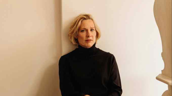 A blonde woman in a black polo neck sits on a wooden step and looks somewhat austere