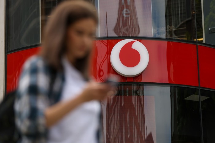 A pedestrian uses a mobile phone as they pass a Vodafone Group Plc mobile phone store