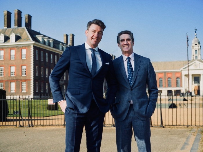 Two men in blue suits smiling outside a grand building on a bright day
