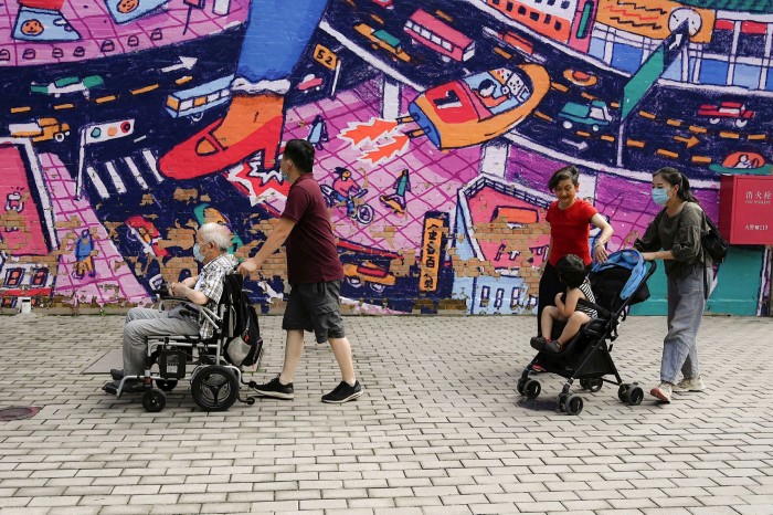 A man pushes a wheelchair with an elderly man and two women push a stroller with a child past a peeling wallpaper at 798 art district in Beijing