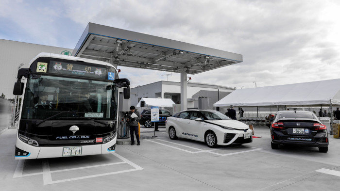 At a hydrogen refueling station, a fuel cell bus and two fuel cell cars are refueling. The bus has “RESERVED” displayed on its sign and features a wheelchair symbol. The station has a sleek, contemporary design with a large canopy overhead. Several people are present, with a tent and additional structures visible in the background
