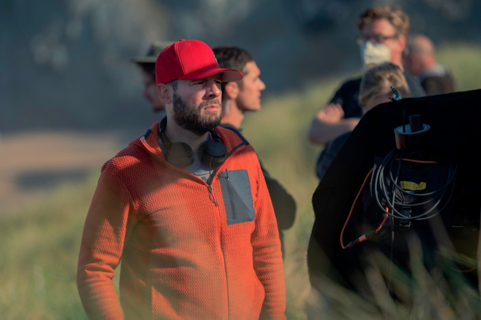 A man in an orange fleece and red baseball cap, with headphones slung around his neck, stands in outside location, with two other crew members behind him