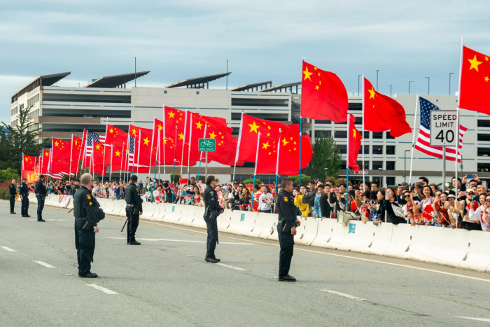 People stand waving flags behind a road block while security men and women with weapons stand on the road. There is a building in the background