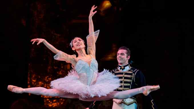 A ballerina with a joyful expression leaps with arms and legs outstretched while a male ballet dancer looks on, smiling