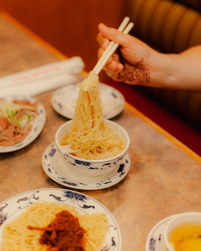 A hand holding a pair of chopsticks taking noodles from a small bowl at Max Noodle House