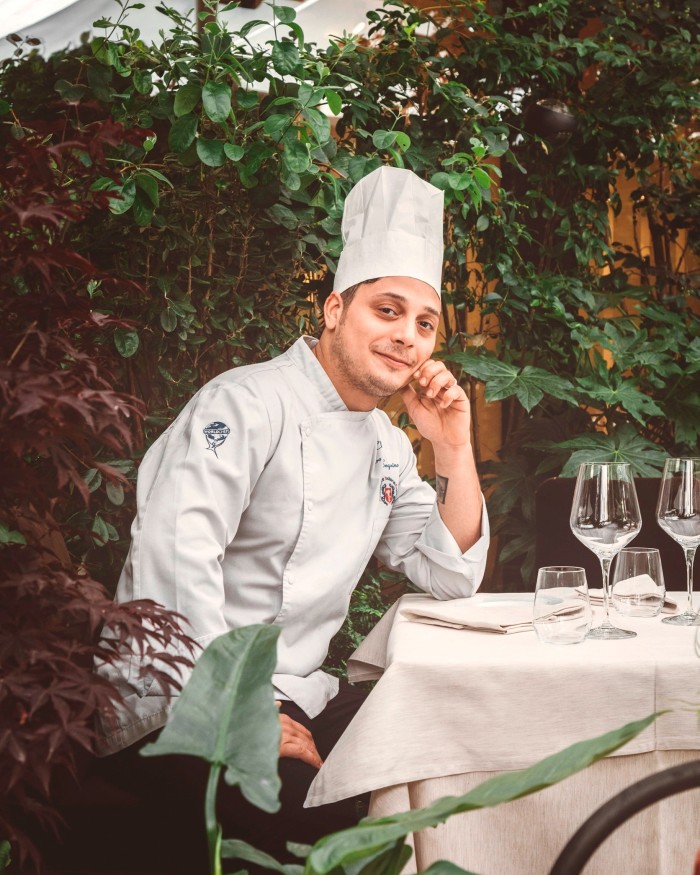 Il Baretto chef Ciro Sequino wearing chef’s whites and a toque and sitting at a garden table in the restaurant, surrounded by foliage