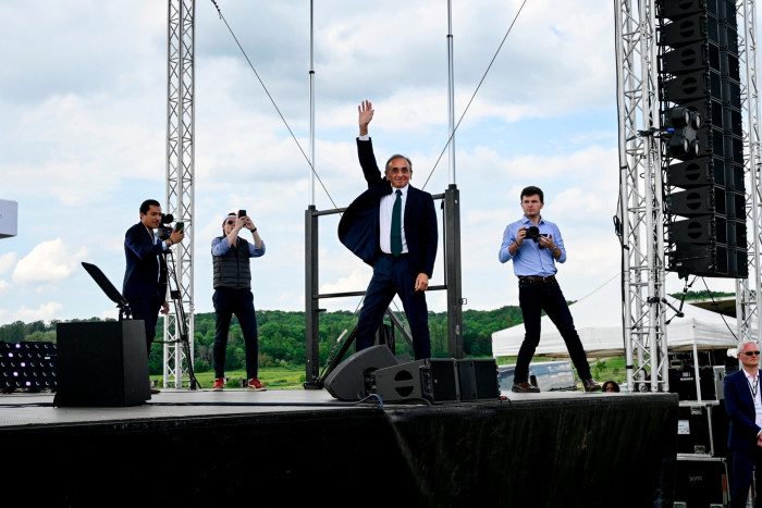 A man wearing a suit stands on a stage with his right arm in the air. Three men are also on stage taking his photograph