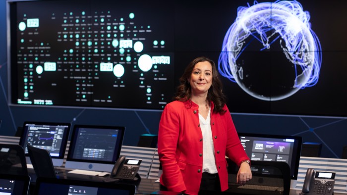 A woman standing in the middle of a room filled with computers. There is a giant monitor behind her showing a globe and other digital info