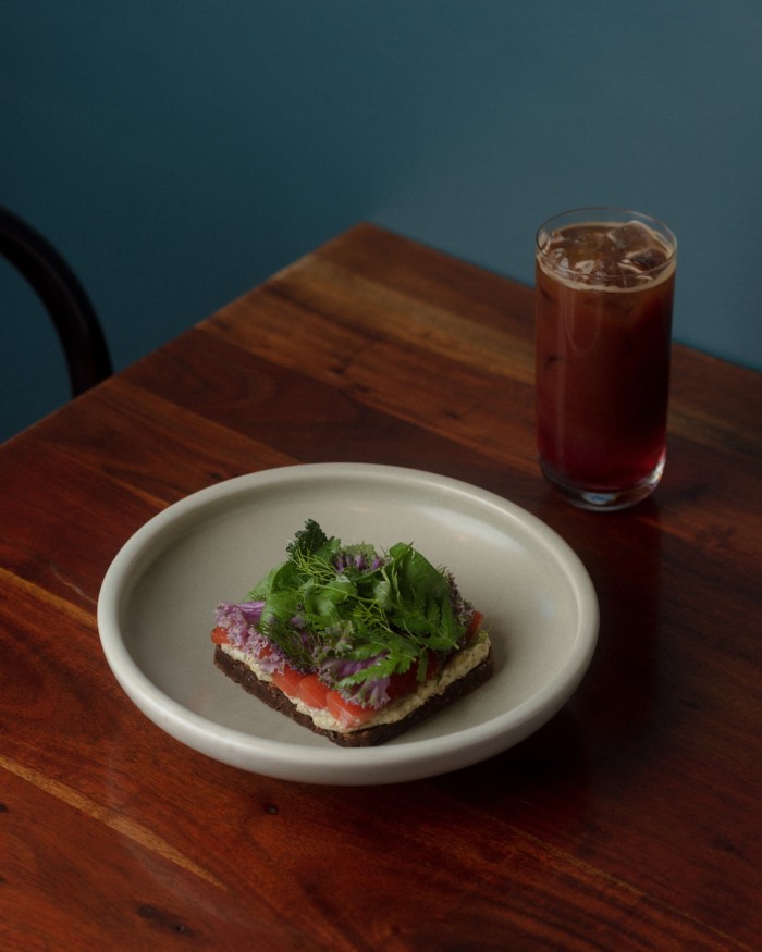 A plate of gravadlax on rye bread with elderflower remoulade, shallot, pea shoots and picked dill and a glass of cold-brew coffee on a table at Novella