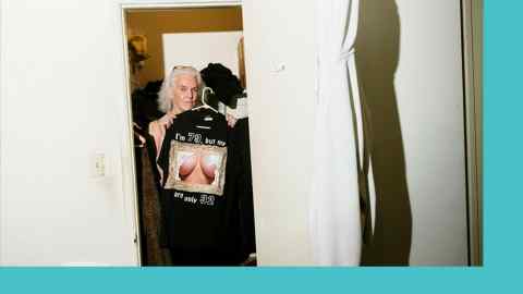A woman seen through a doorway holding up a T-shirt which says, “I’m 79 but my [picture of breasts] are only 32”