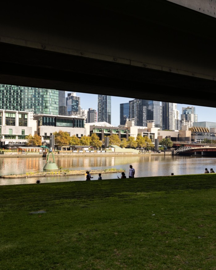 Looking out from beneath a bridge at lawn in Enterprize Park, leading down to the banks of the Yarra, where people are sitting