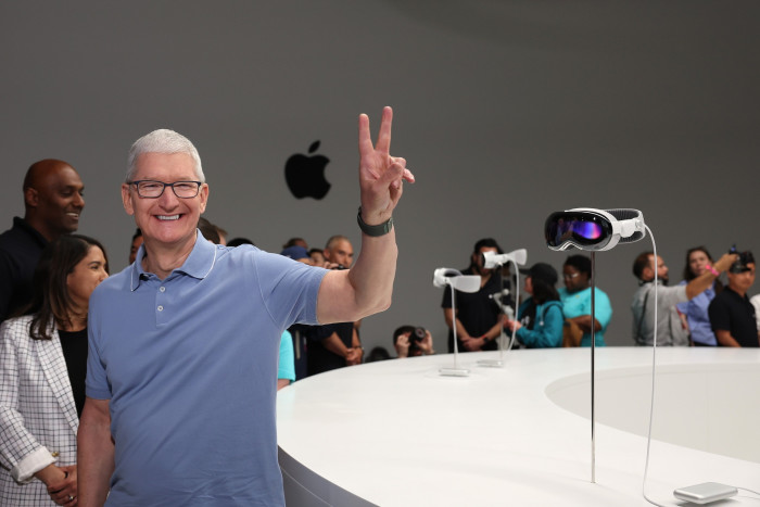 Tim Cook holds up one hand in a V for victory sign while standing next to a Vision Pro headset on display
