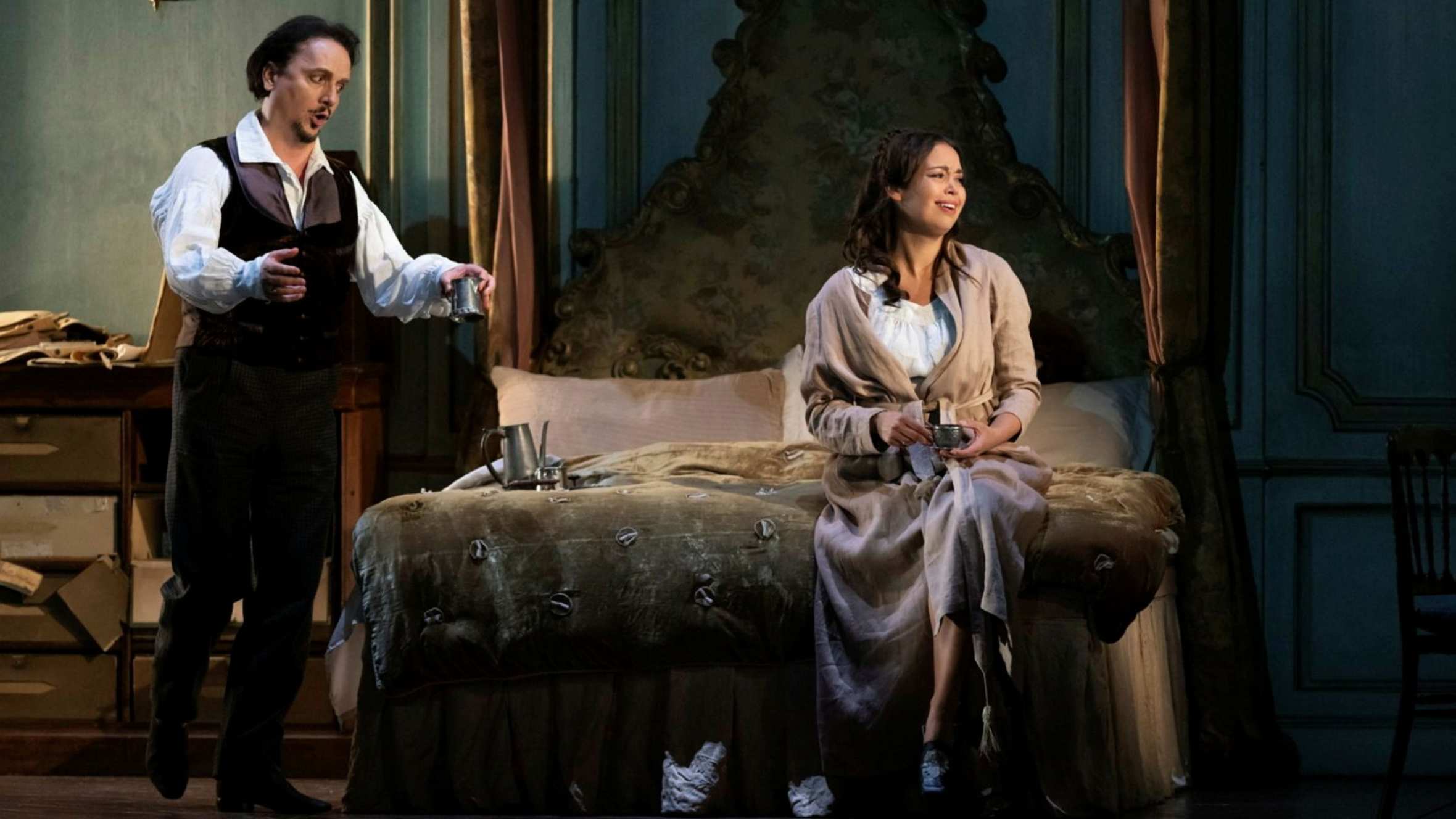 A man and a woman wearing 19th-century costumes are in a bedroom; the man stands singing passionately to the woman, who sits on the bed with her head turned away from him