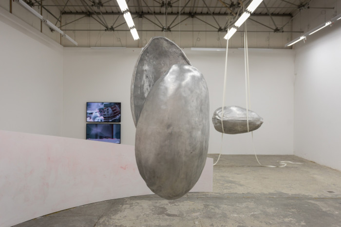 A huge, silver sculpture of a pistachio shell hangs from the ceiling of a gallery. Behind it, a smaller shell floats horizontally to its right while two screens are visible on its left