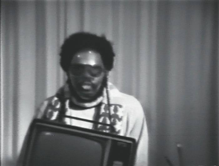 A blurry black and white still of a man wearing sunglasses behind a TV