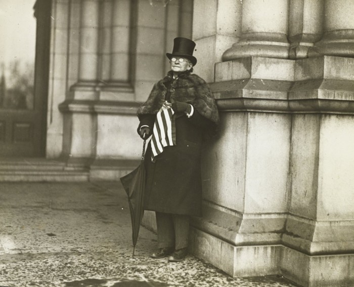 A figure wearing a top hat and greatcoat stands next to the pillar of a large building, holding a US flag
